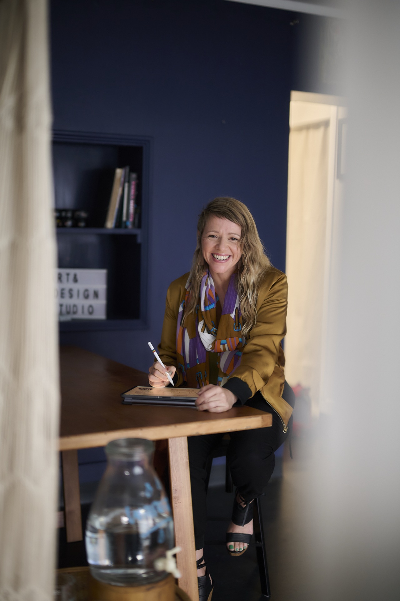 Megan Salole sitting at a desk with an Apple Pencil, smiling, in a navy blue studio
